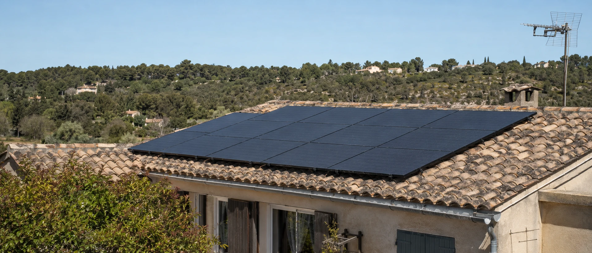 Toiture de pavillon en tuiles canal vue en légère plongée : champ de modules photovoltaïques noirs posés sur rails, cheminée crépie et collines boisées en arrière-plan.